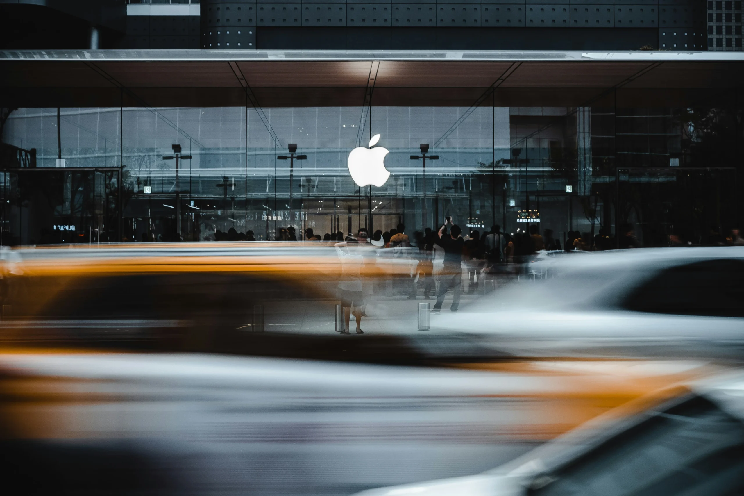 A picture of the white apple logo on the outside of an apple store while cars are blurred in the foreground. 