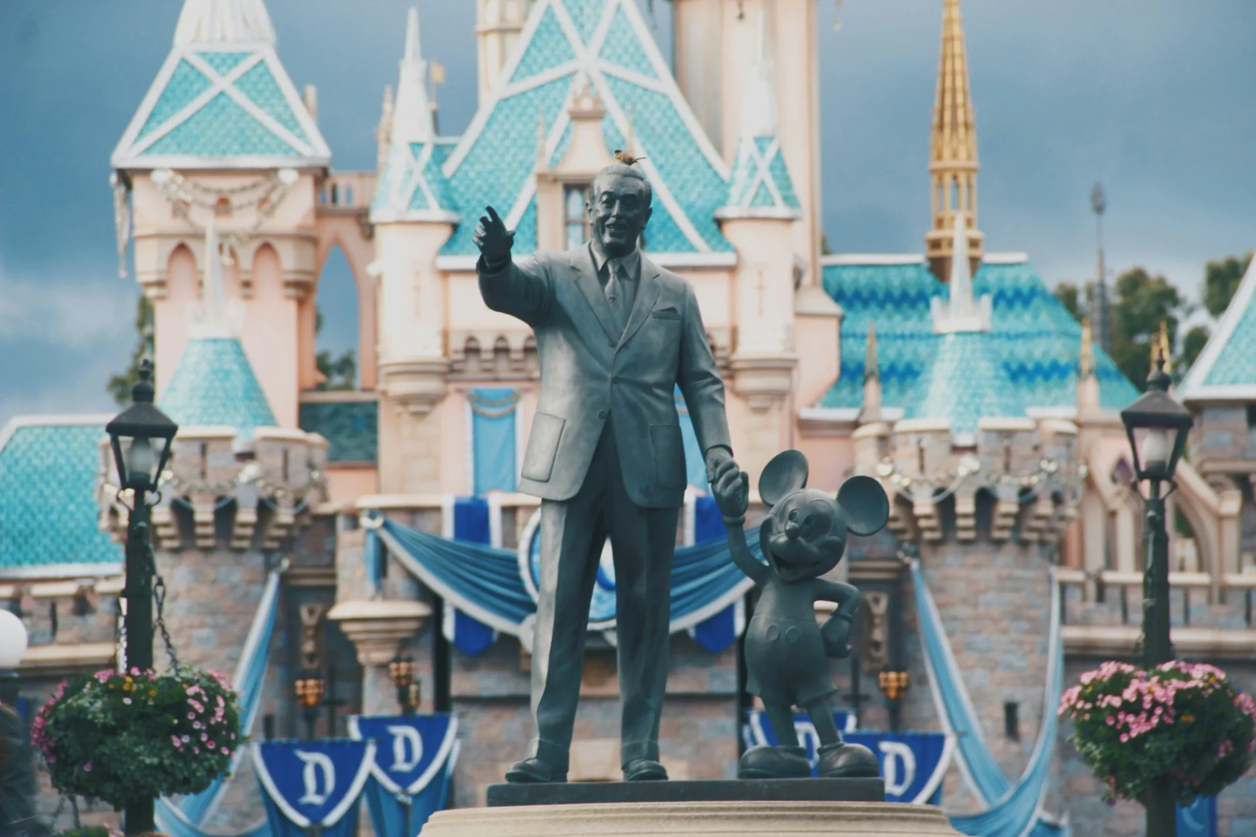 A teal statue of Walt Disney holding mickey mouse's hand in front of the Disney castle that represents how Disney is an inspirational brand.